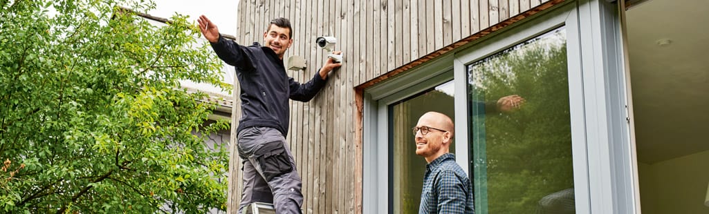 Ein Mann steht auf einer Leiter und montiert gerade eine Überwachungskamera an einer Hauswand. Der Mann zeigt einem anderen Mann, der lächelnd auf dem Boden steht, mittels Handzeichen den Bereich, der von der Kamera erfasst wird.