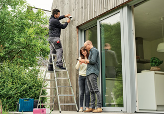 Ein Mann steht auf einer Leiter und montiert eine Überwachungskamera an der Außenwand eines Hauses. Neben ihm auf dem Boden steht ein Päärchen, das gemeinsam fröhlich in ein Smartphone schaut. 