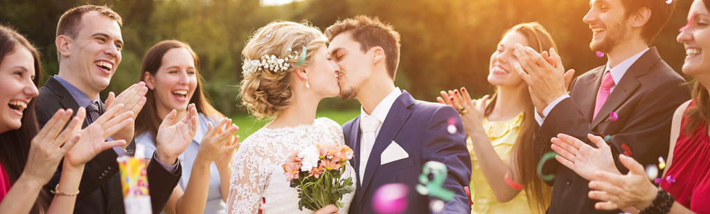 Ein Hochzeitspaar steht in der Mitte von lachenden und klatschenden Menschen und küsst sich.