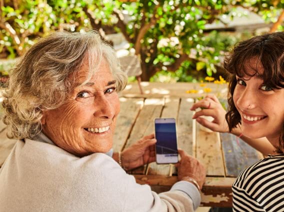 Eine ältere Frau und eine jüngere Frau schauen über Ihre schulter in die Kamera und lächeln. In der Hand hält die ältere Frau ein Handy.
